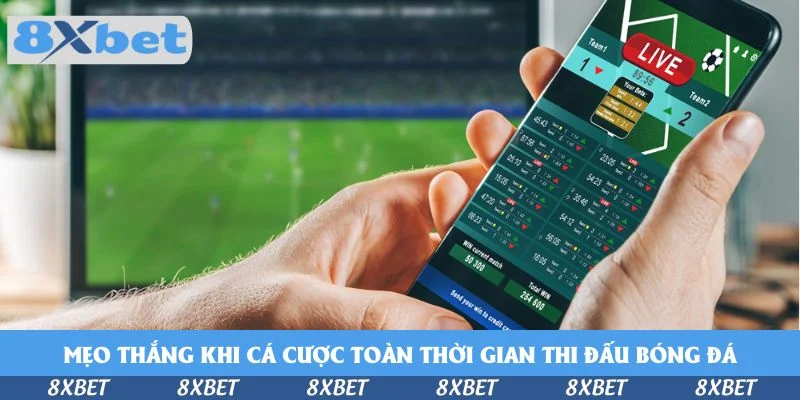 Tay cầm điện thoại mở giao diện cược bóng đá trực tiếp có nhãn LIVE trước màn hình trận đấu