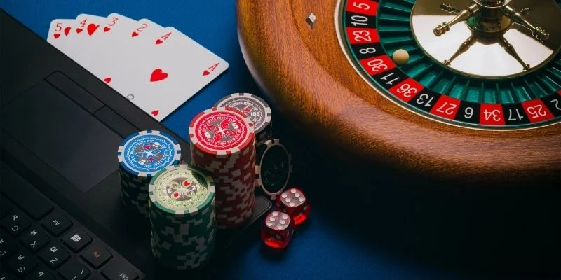 Phỉnh casino, quân bài, xúc xắc và bàn quay roulette đặt cạnh bàn phím laptop